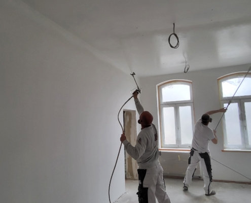 Zwei Malergesellen in weißer Arbeitskleidung streichen bzw. sprühen die Decke eines Raumes. Der Raum ist leer und im Rohbauzustand, mit unverkleidetem Boden und freiliegenden Kabeln an der Decke. Sonnenlicht fällt durch zwei große Fenster auf der rechten Seite.