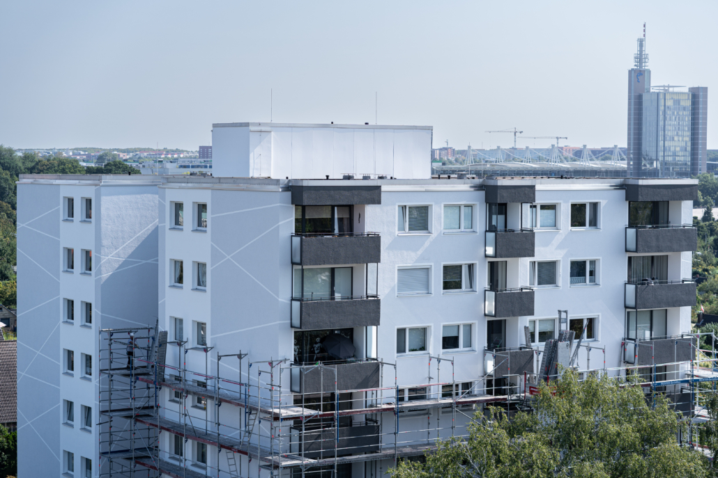 Mehrstöckiges Wohngebäude mit frisch renovierter Fassade in Weiß und dunklen Balkonverkleidungen. Die Fassade ist mit hellen diagonalen Linien gestaltet. Vor dem Gebäude ist ein Baugerüst aufgebaut, was auf laufende Fassadensanierung und Wärmedämmung hinweist. Im Hintergrund sind Bäume, ein Stadion mit Seilspannkonstruktion und moderne Bürogebäude zu sehen. Der Himmel ist klar und hellblau.