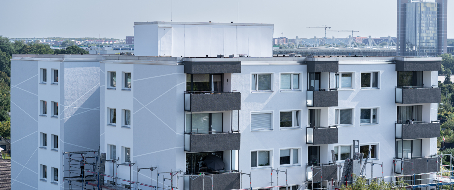 Mehrstöckiges Wohngebäude mit frisch renovierter Fassade in Weiß und dunklen Balkonverkleidungen. Die Fassade ist mit hellen diagonalen Linien gestaltet. Vor dem Gebäude ist ein Baugerüst aufgebaut, was auf laufende Fassadensanierung und Wärmedämmung hinweist. Im Hintergrund sind Bäume, ein Stadion mit Seilspannkonstruktion und moderne Bürogebäude zu sehen. Der Himmel ist klar und hellblau.