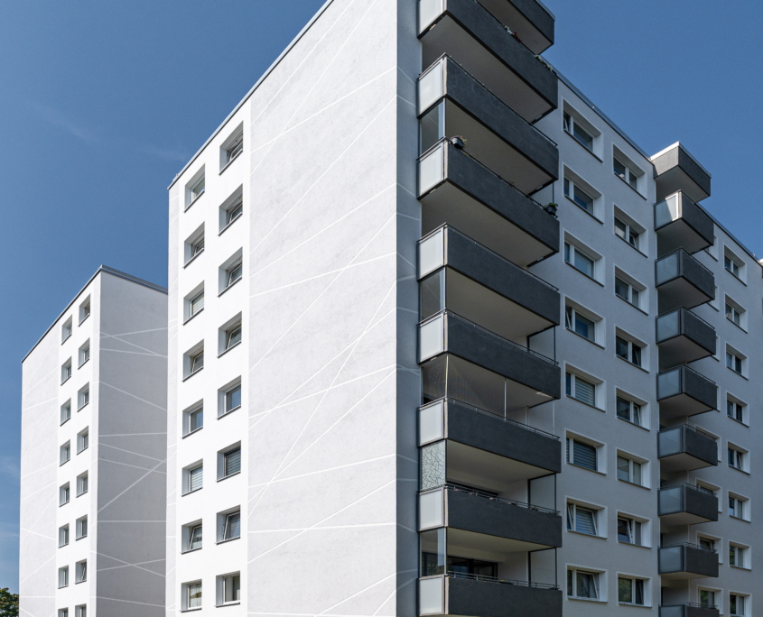 Moderne, frisch renovierte Wohngebäude-Fassade mit weiß geputzter Wärmedämmung und dekorativen diagonalen Linien. Die Gebäude haben mehrere Stockwerke und Balkone mit Glasbrüstungen und Blumen. Im Hintergrund sind weitere Gebäude, Bäume und ein leicht bewölkter blauer Himmel zu sehen.