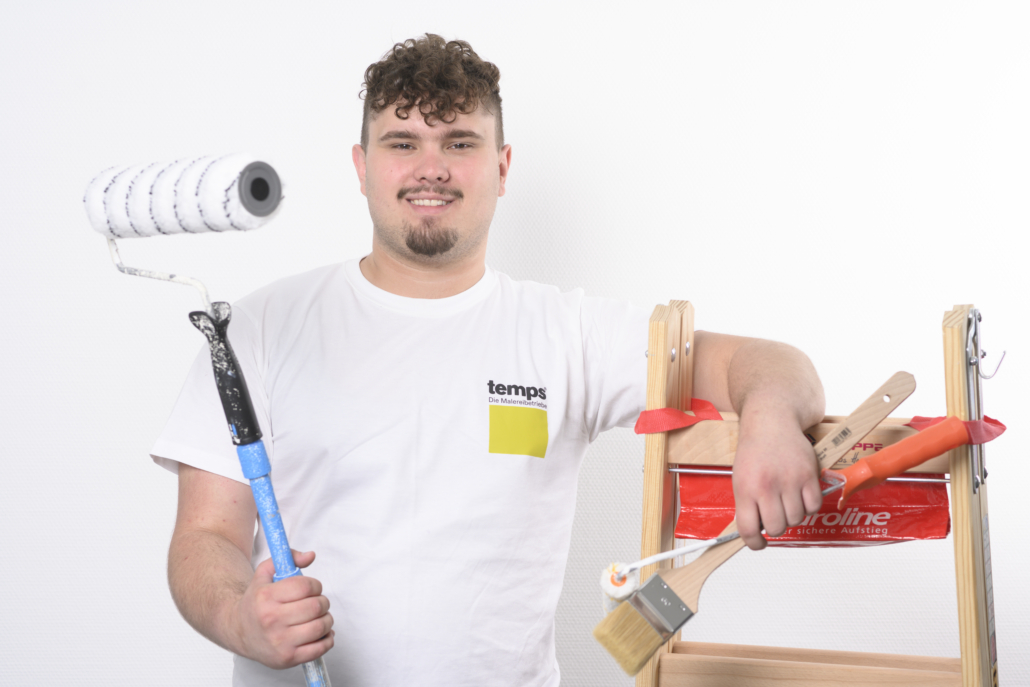 Ein weiß gekleideter Handwerker lehnt mit Pinsel und Rolle in den Hand an einer Holzleiter und grinst selbstbewusst in die Kamera - so wird Malerhandwerk erlebbar