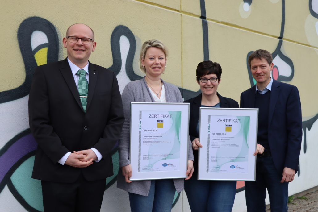 Drei temps-Verantwortliche stehen nebeneinander vor einer bunten Wand mit temps-Graffiti, schauen lächelnd in die Kamera und freuen sich über die im Rahmen der Zertifizierung erhaltenen Urkunden, übergeben durch den DEKRA-Vertriebsleiter.