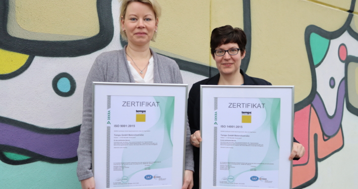 Zwei Büro-Mitarbeiterinnen stehen mit den Zertifizierungen in der Hand vor einer bunten Mauer mit temps-Schriftzug