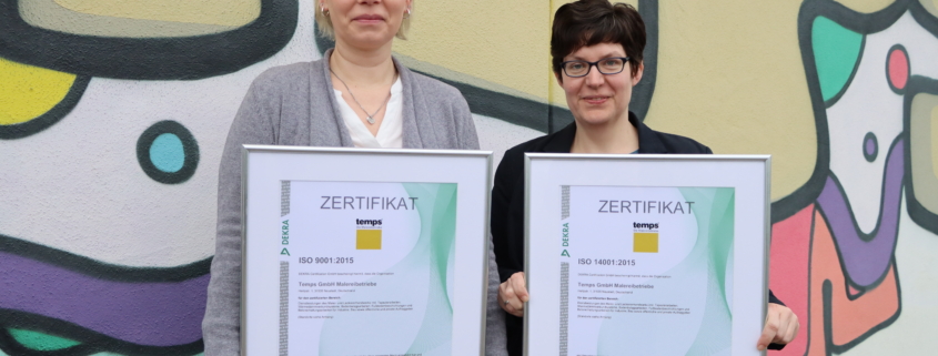 Zwei Büro-Mitarbeiterinnen stehen mit den Zertifizierungen in der Hand vor einer bunten Mauer mit temps-Schriftzug